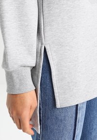 Felpa grigia con polsini a costine, spacchi laterali e vestibilità rilassata, abbinata a jeans in denim blu. Il tessuto sembra morbido e lavorato.