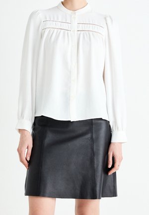 Femme portant un chemisier blanc à manches longues avec des détails en dentelle et une jupe trapèze en cuir noir.