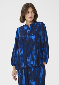 Laivansininen paita, jossa on abstrakteja sinisiä ja mustia kuvioita. Varustettu pyöreällä kaula-aukolla, nappilistan kanssa ja elastisilla hihansuilla rentoa istuvuutta varten.