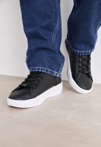 Svarta läderskor med strukturerad yta, vit gummisula och traditionell snörningsdesign, parat med blå denimjeans.