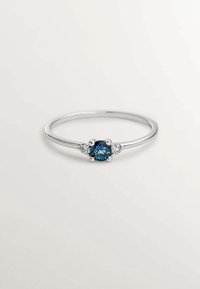Anillo de plata con una gema azul central flanqueada por dos piedras claras más pequeñas, con una banda delgada y elegante y un acabado pulido.
