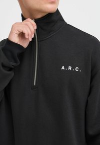 Hombre subiendo la cremallera de un sudadera negra de media cremallera con el texto blanco "A.R.C." en el pecho, mostrando la mano, el cuello y parte de la cara sobre un fondo neutro.