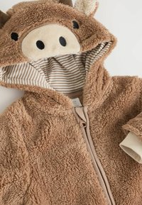 Fleece-Einteiler mit Tiermotiven in Brauntönen, ausgestattet mit einer strukturierten Außenseite, gestreiftem Innenfutter und einem Reißverschluss. Mit Kuhmotiven und Ohren.