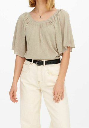 Kvinde iført en beige bluse med rund hals og flagrende ærmer, stoppet ned i højtaljede off-white bukser med et sort bælte.