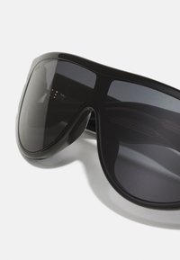 Occhiali da sole neri oversize con una montatura in plastica lucida e liscia e lenti scure, caratterizzati da un design avvolgente.