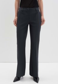 Pantalon gris foncé sur mesure avec une coupe droite, tissu lisse, passants de ceinture et plis sur le devant, associé à des escarpins noirs à bout pointu.
