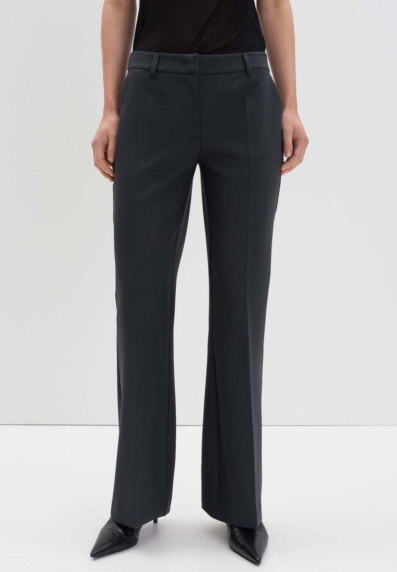 Pantalon gris foncé sur mesure avec une coupe droite, tissu lisse, passants de ceinture et plis sur le devant, associé à des escarpins noirs à bout pointu.