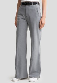 Pantalons gris sur mesure avec jambes larges, plis à l'avant et une ceinture noire. Le tissu a une texture lisse. Portés avec des sneakers blancs.