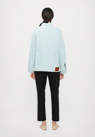 Light blue denim jacket with a casual fit, featuring a back yoke, side slits, and brown leather label at the hem, paired with black trousers.