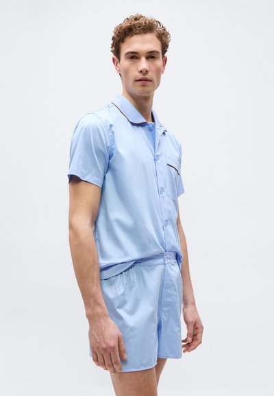 Νέος άνδρας που στέκεται φορώντας ανοιχτό μπλε πουκάμισο με κοντά μανίκια και κουμπιά, μαζί με κοντά παντελόνια που ταιριάζουν, με καφέ περιθώριο στο γιακά και την τσέπη.