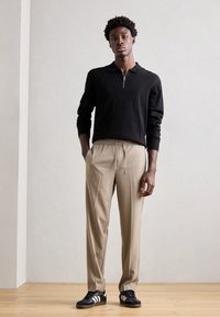 Polo noir à manches longues avec col zippé, associé à un pantalon beige avec taille à cordon, et des baskets noires avec des accents blancs.