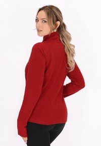 Rdeča flis majica z dolgimi rokavi, visoko ovratnikom, prilegajočega kroja in mehkim materialom. Opazne šive, kombinirana z črnimi leggins.