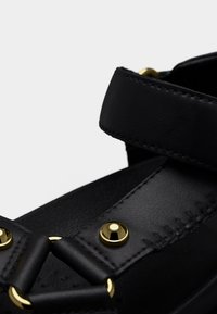 Primer plano de una sandalia de cuero negro que muestra tiras texturizadas con tachuelas y anillos de metal en tono dorado.