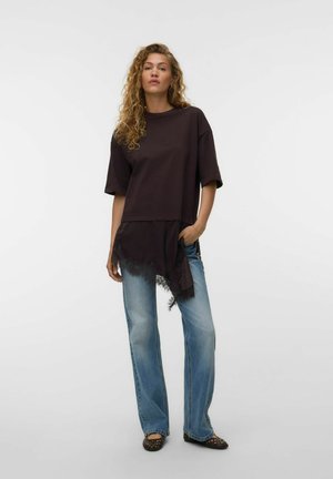 Femme aux cheveux bouclés portant un haut noir ample avec un détail en dentelle, un jean bleu clair et des chaussures noires ouvertes à l'avant, debout sur un fond blanc.