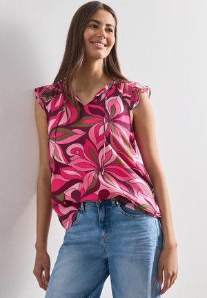 Mujer sonriendo con una blusa sin mangas de flores rosas y rojas con hombros con volantes y vaqueros azul claro sobre un fondo claro y liso.