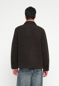 Braune strukturierte Jacke mit Kragen, langen Ärmeln und geknöpften Manschetten, kombiniert mit hellblauen Denimhosen mit großen Taschen.