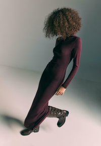 Dlhé bordové šaty s vysokým výstrihom a dlhými rukávmi, doplnené hnedými koženými čižmami po kolená s prackami a textúrovaným povrchom.