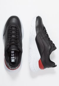 Baskets en cuir noir avec une semelle blanche mouchetée, des accents rouges et un design à lacets. Le logo est visible sur la languette et le talon.