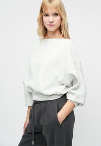 Svijetlosiva, oversized dukserica s brodskim izrezom i rebrastim rubom. Mekana tekstura, u kombinaciji s tamnosivim, širokim duksericama.