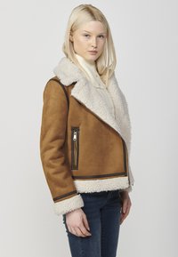 Giacca in faux suede marrone con colletto e rifiniture in shearling crema, dotata di una tasca diagonale con zip e una texture morbida. Design edge-to-edge.
