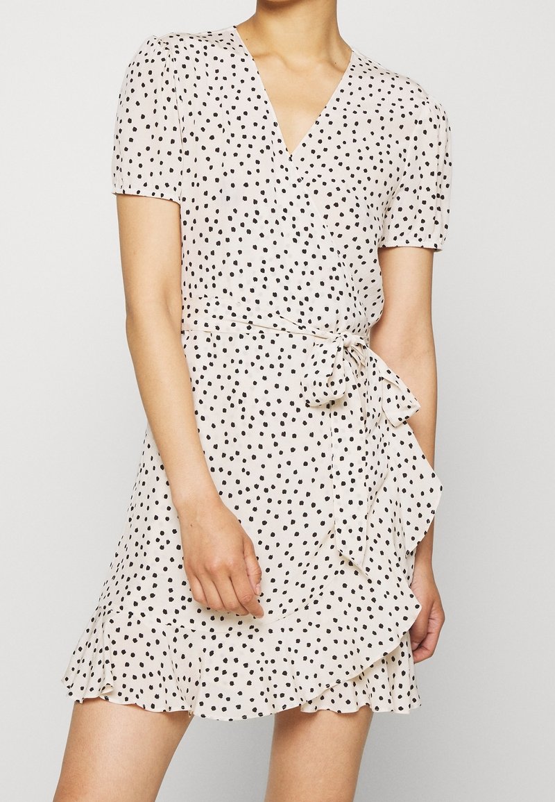 Femme portant une robe portefeuille claire à manches courtes à pois noirs, ceinture nouée à la taille et ourlet à volants, debout devant un fond uni.