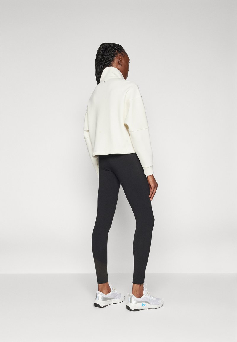 Donna con capelli intrecciati, che indossa un felpa bianca a collo alto, leggings neri e scarpe da ginnastica bianche, in piedi su uno sfondo leggero e uniforme.