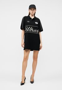 Mujer con vestido polo negro de gran tamaño con el texto "Ultras", gorra negra y zapatos de tacón negros, de pie contra un fondo liso.