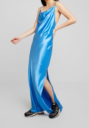 Femme portant une robe sans manches en satin bleu brillant avec une fente haute sur la cuisse, associée à des baskets noires Nike, sur un fond gris.