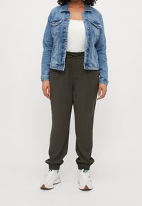Veste en denim bleu clair avec fermeture à boutons et poches, associée à un jogging vert olive avec taille élastique et poignets. Baskets blanches.