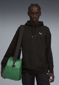 Sudadera negra con bolsillo frontal, que presenta un logo blanco; la modelo sostiene un bolso de cuero verde con una textura suave y correa ajustable.