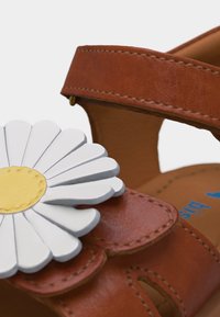Καφέ δερμάτινο σανδάλι με μεγάλο λευκό και κίτρινο διακοσμητικό μαργαρίτας στο λουράκι, με εμφανή ραφές και σχεδιασμό με κοψίματα.
