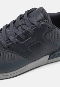 Zapatilla deportiva de color gris oscuro que presenta una combinación de cuero liso y malla transpirable, con cordones y una marca en el lateral. Presenta perforaciones sutiles.
