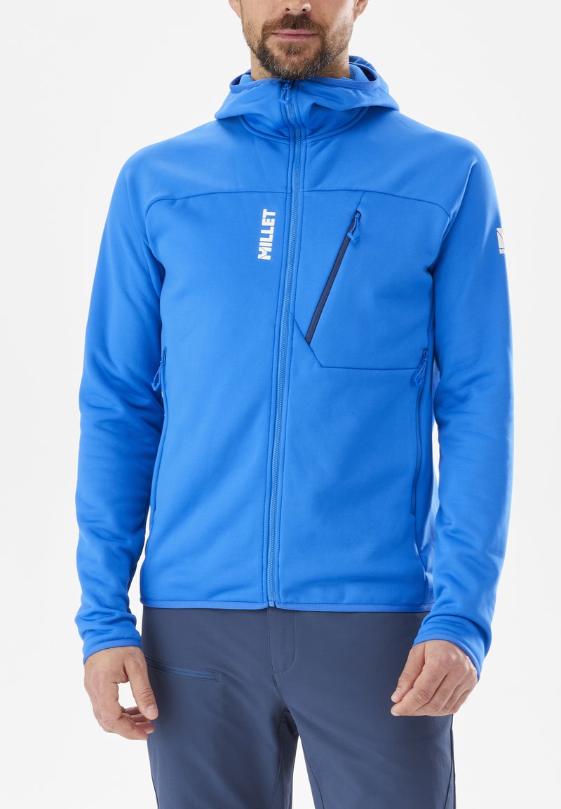 Homme portant une veste à capuche zippée bleu vif avec des poches zippées et un pantalon bleu foncé, se tenant devant un fond blanc uni.