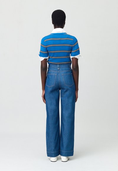 Chemise en tricot côtelé rayée bleue avec col blanc, associée à un jean en denim bleu à jambe large taille haute. Le mannequin tourne le dos à l'appareil photo.