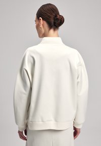 Felpa oversize bianca con polsini e colletto a coste, caratterizzata da una texture liscia e un design minimalista. La vista posteriore evidenzia la semplicità.