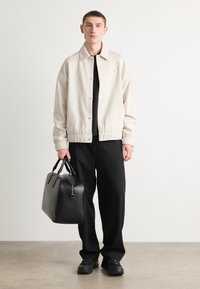 Giacca bomber beige chiaro con colletto, t-shirt nera sotto, pantaloni neri e borsa in pelle nera con due manici.