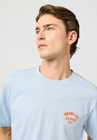 Ljusblå bomullströja med en broderad "WRANGLER JEANS 1947"-logotyp i orange på vänster bröstområde. Klassisk crew neck-design.