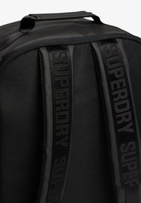 Zwarte rugzak van glad nylonmateriaal, opvallende "SUPERDRY" branding op de banden en een gestructureerde rechthoekige vorm met een handvat aan de bovenkant.