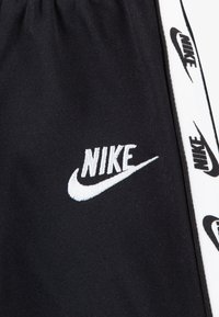 Fekete sportnadrág elasztikus derékpánttal, fehér hímzett Nike logóval és oldalsó csíkokkal, amelyeken ismétlődő logóminták láthatók.