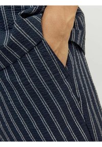 Tamsiai mėlyna apranga su tekstūruotu audiniu, pasižyminti baltais vertikaliais dryžiais, turi šoninę kišenę ir sulankstytą apykaklės detalę.