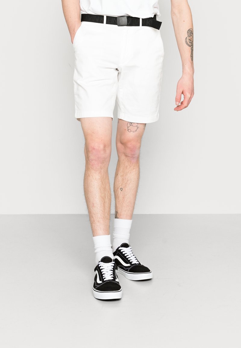 Shorts bianchi in cotone con tasche laterali, abbinati a sneakers nere e bianche a basso profilo e calze bianche. Presentano una cintura nera.