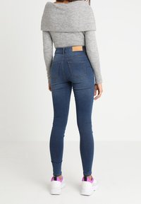 Jeans blu skinny con vita alta, tasche posteriori e un'etichetta in pelle beige. Abbinati a un maglione grigio scollato sulle spalle e sneakers bianche.