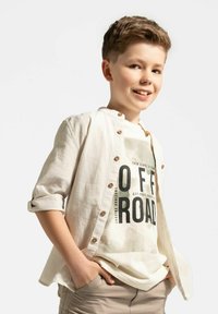 Jasno beżowa lniana koszula z podwiniętymi rękawami i drewnianymi guzikami, noszona na białym t-shircie z grafiką, na którym widnieje wyraźny czarny tekst, w luźnym kroju.