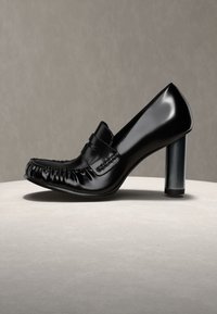 Mocassin à talon haut en cuir noir brillant, avec bout arrondi et détails froncés, présenté sur une surface claire avec un fond gris.