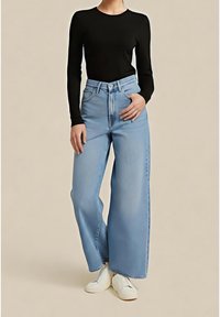 Personne portant un jean taille haute à jambes larges bleu clair, un haut noir moulant à manches longues et des chaussures blanches, debout avec une main dans la poche.