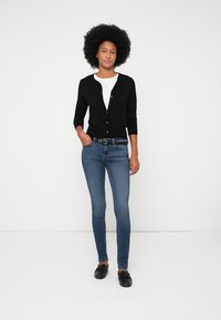 Juodas kardiganas su apvaliu iškirpimu ir sagčių uždarymu, derinamas su baltu marškinėliu, siaurais mėlynais džinsais ir juodais loaferiais.