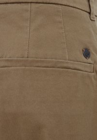 Smeđa pamučna tkanina ima teksturiranu površinu s dizajnom džepa i tamnom okruglom logotip oznakom sa strane.