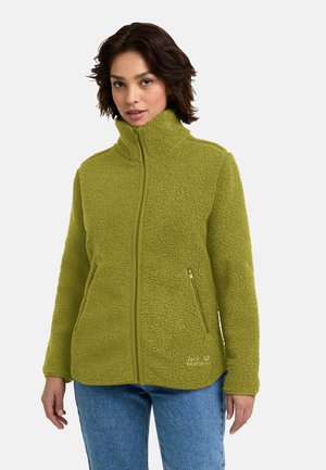 Vrouw draagt een groene fleecejas met ritssluiting, hoge kraag en twee zijritszakken, gecombineerd met een blauwe spijkerbroek.