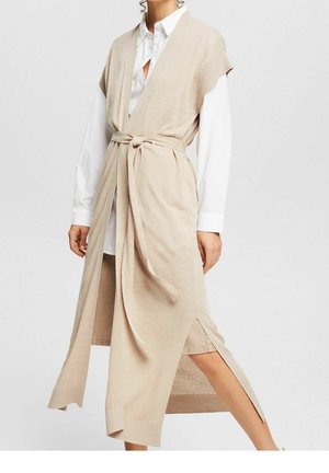 Beige knitted wrap dress with short sleeves, tie waist, and side slits, layered over a white button-up shirt. Soft texture and relaxed fit.