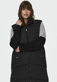 Svart pufferväst med hög krage, grå tröjliknande ärmar, dragkedjestängning och sidofickor. Kviltad textur och kontrasterande material.